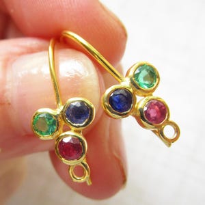 May include: A pair of gold-tone earrings featuring a cluster of three round faceted gemstones in green, blue, and red. Each earring has a curved wire hook for attachment and a small circular loop at the bottom for hanging additional jewellery components.