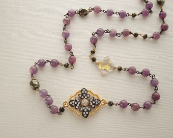 May include: A purple and gold beaded necklace with a black and white flower pendant. The pendant has a clear stone in the center.