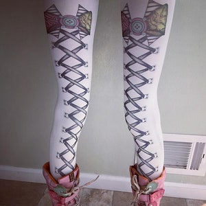 Peut inclure: Collants blancs avec un motif de corset noir et gris et un nœud rose avec un bouton sur chaque jambe. Les collants sont associés à des bottes à lacets roses et blanches.
