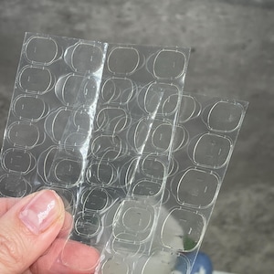 May include: A sheet of clear adhesive tabs for attaching false nails. The tabs are arranged in a grid pattern on a clear plastic sheet.
