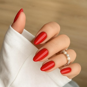 Peut inclure: Gros plan sur une main avec de longs ongles en amande peints en rouge vif. Une bague en argent avec des perles orne l'annulaire. La main est partiellement couverte par une chemise blanche à manches longues.