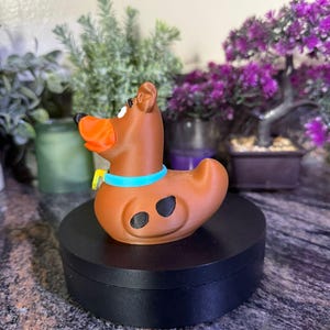 May include: A brown dog-shaped rubber duck figure with a bright orange snout, a light blue collar with a yellow tag, and two black spots on its side. The figure is positioned in profile, displaying a smooth, matte finish and a curved tail.