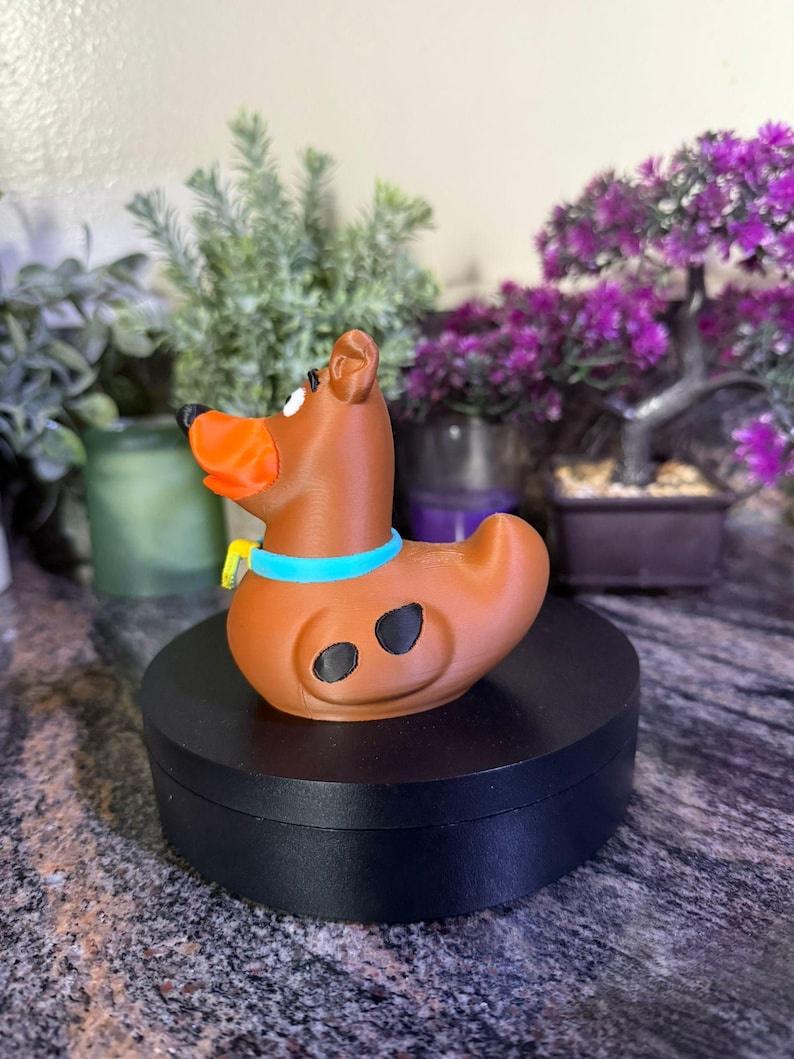 May include: A brown dog-shaped rubber duck figure with a bright orange snout, a light blue collar with a yellow tag, and two black spots on its side. The figure is positioned in profile, displaying a smooth, matte finish and a curved tail.