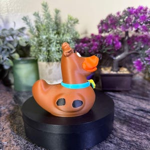 May include: A brown rubber duck-shaped figure designed to resemble a dog, featuring an orange snout, a bright blue collar with a small yellow tag, and two black oval spots on its side. The figure has a smooth, molded surface and a distinct duck-like tail.