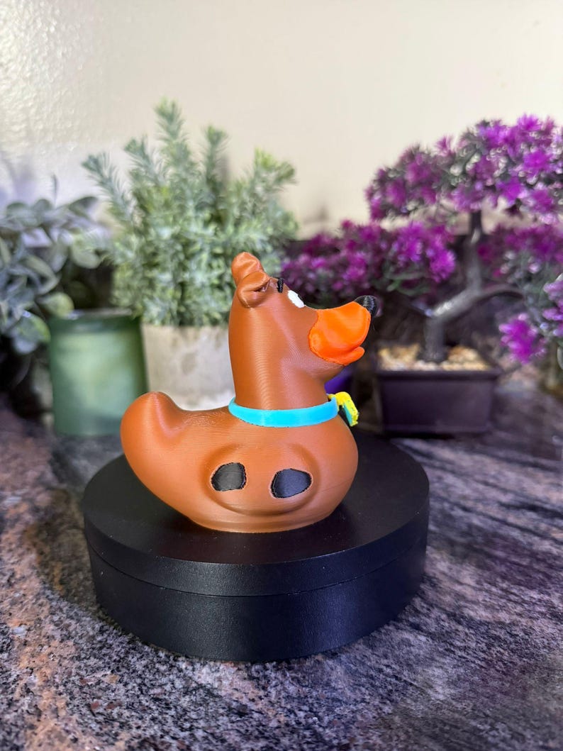 May include: A brown rubber duck-shaped figure designed to resemble a dog, featuring an orange snout, a bright blue collar with a small yellow tag, and two black oval spots on its side. The figure has a smooth, molded surface and a distinct duck-like tail.