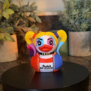 Op de afbeelding: Een decoratieve badeend met een Harley Quinn-ge&iuml;nspireerd ontwerp. De eend heeft een wit gezicht met blauwe en roze make-up, gele staartjes en een rood en blauw outfit. De tekst "Daddy's Lil' Kooki" is op de voorkant gedrukt.
