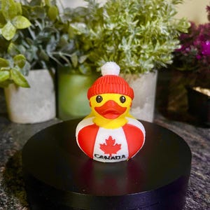 May include: A yellow rubber duck wearing a red and white hat and a red and white life preserver. The life preserver has a maple leaf and the word "CANADA" printed on it. The duck is on a black stand.
