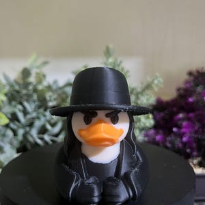 May include: A novelty rubber ducky figurine, dressed in a black hat and coat, with long black hair and an orange beak. The duck has a stern expression. The figurine is on a black stand, with greenery in the background.