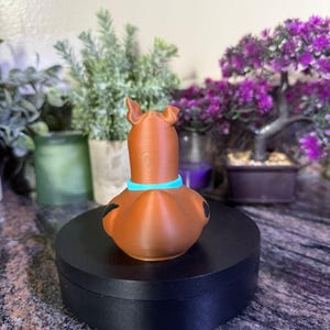 May include: A brown, dog-shaped figurine with a light blue collar around its neck. The figure has two upright ears, a smooth surface texture, and small black spots on its sides. It is positioned on a circular black base.