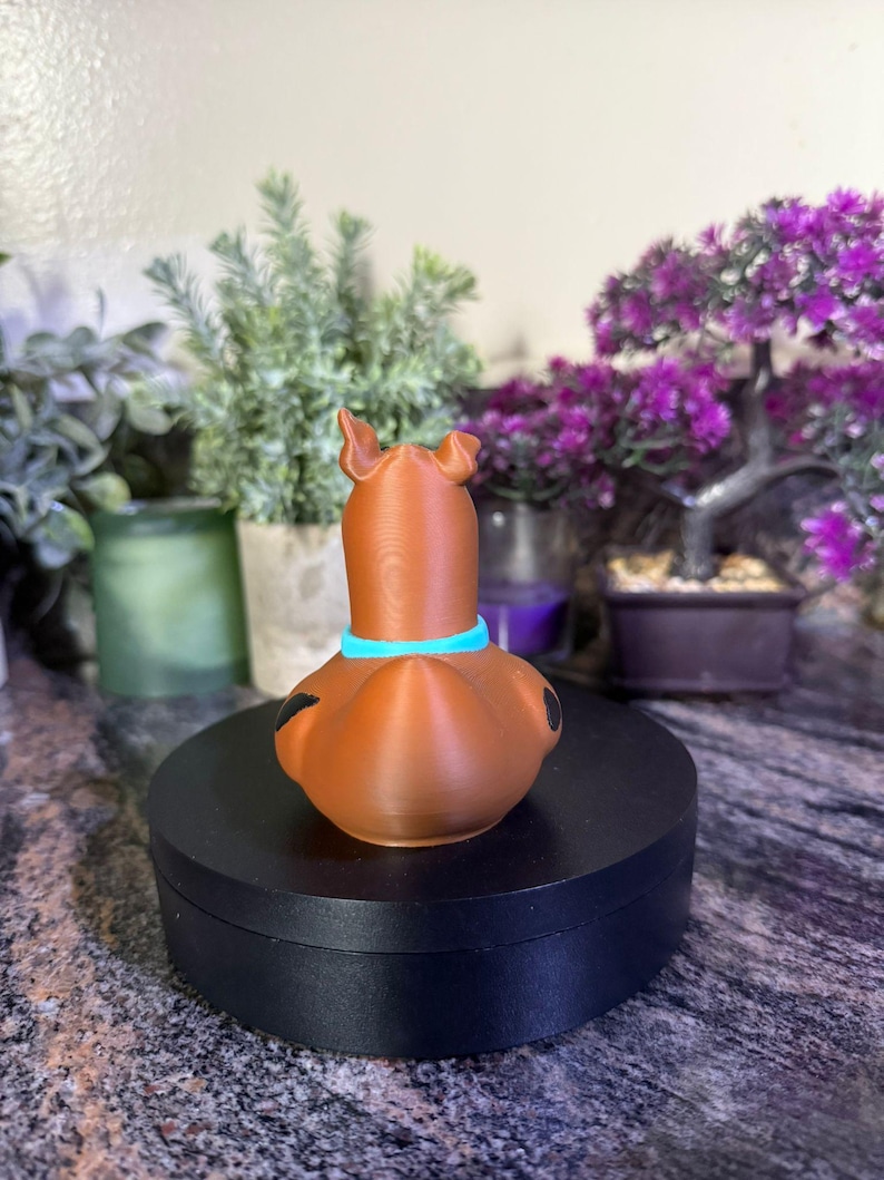 May include: A brown, dog-shaped figurine with a light blue collar around its neck. The figure has two upright ears, a smooth surface texture, and small black spots on its sides. It is positioned on a circular black base.