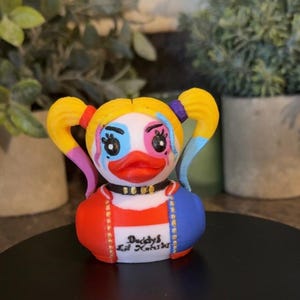 Op de afbeelding: Een nieuwigheid eendenbeeldje met een geschilderd gezicht en kostuum, met gele staartjes, blauwe en roze oogmake-up en een rood-blauwe jas. De tekst "Daddy's Lil' Kooki" staat op de voorkant.