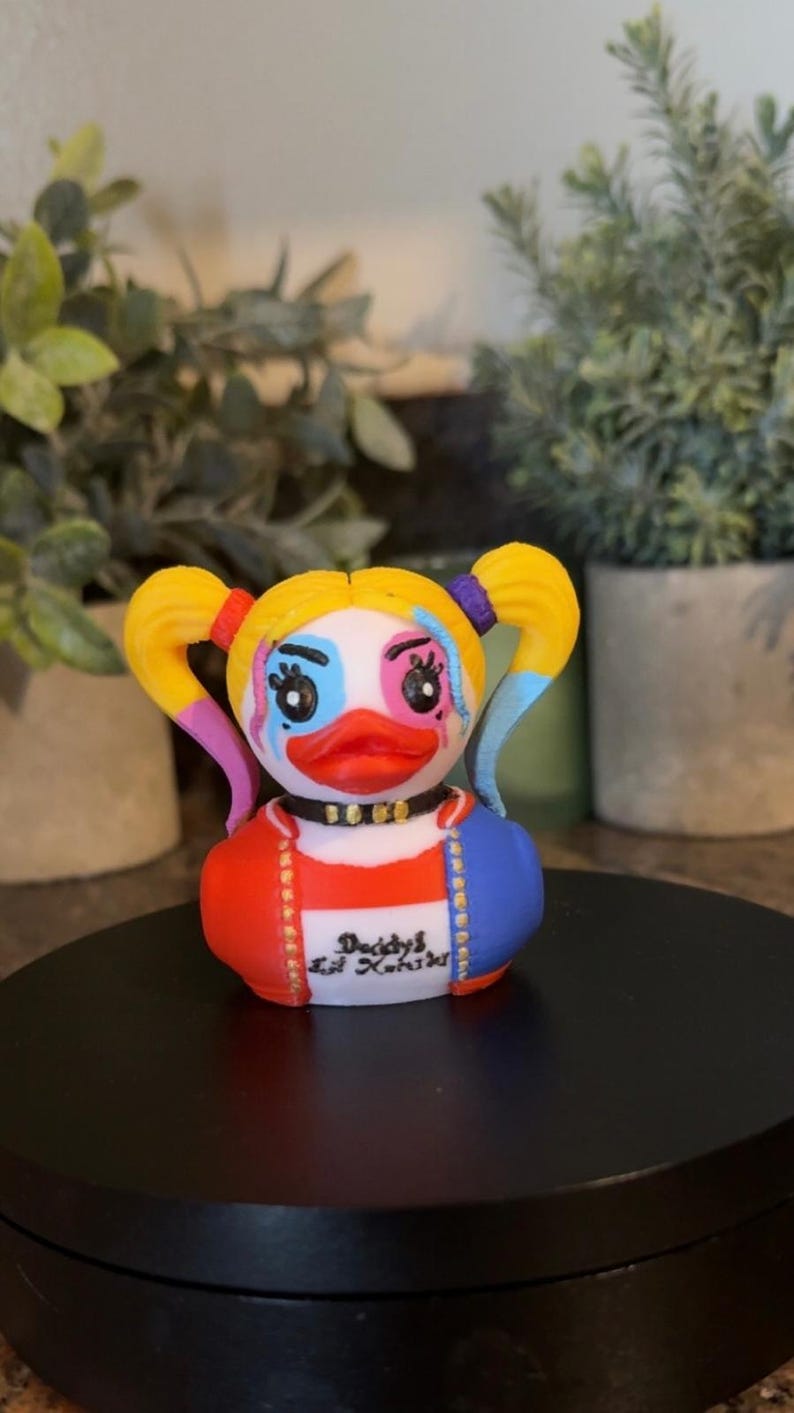 Op de afbeelding: Een nieuwigheid eendenbeeldje met een geschilderd gezicht en kostuum, met gele staartjes, blauwe en roze oogmake-up en een rood-blauwe jas. De tekst "Daddy's Lil' Kooki" staat op de voorkant.