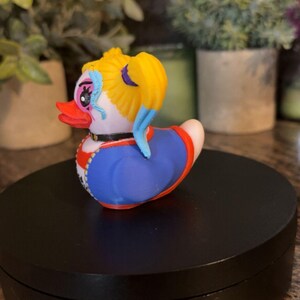Op de afbeelding: Een decoratieve rubberen eend met een uniek ontwerp. De eend heeft een gele haarstijl, blauwe accenten en een blauwe en rode outfit. Hij heeft een zwarte kraag en een rode snavel. De eend staat op een zwart oppervlak.