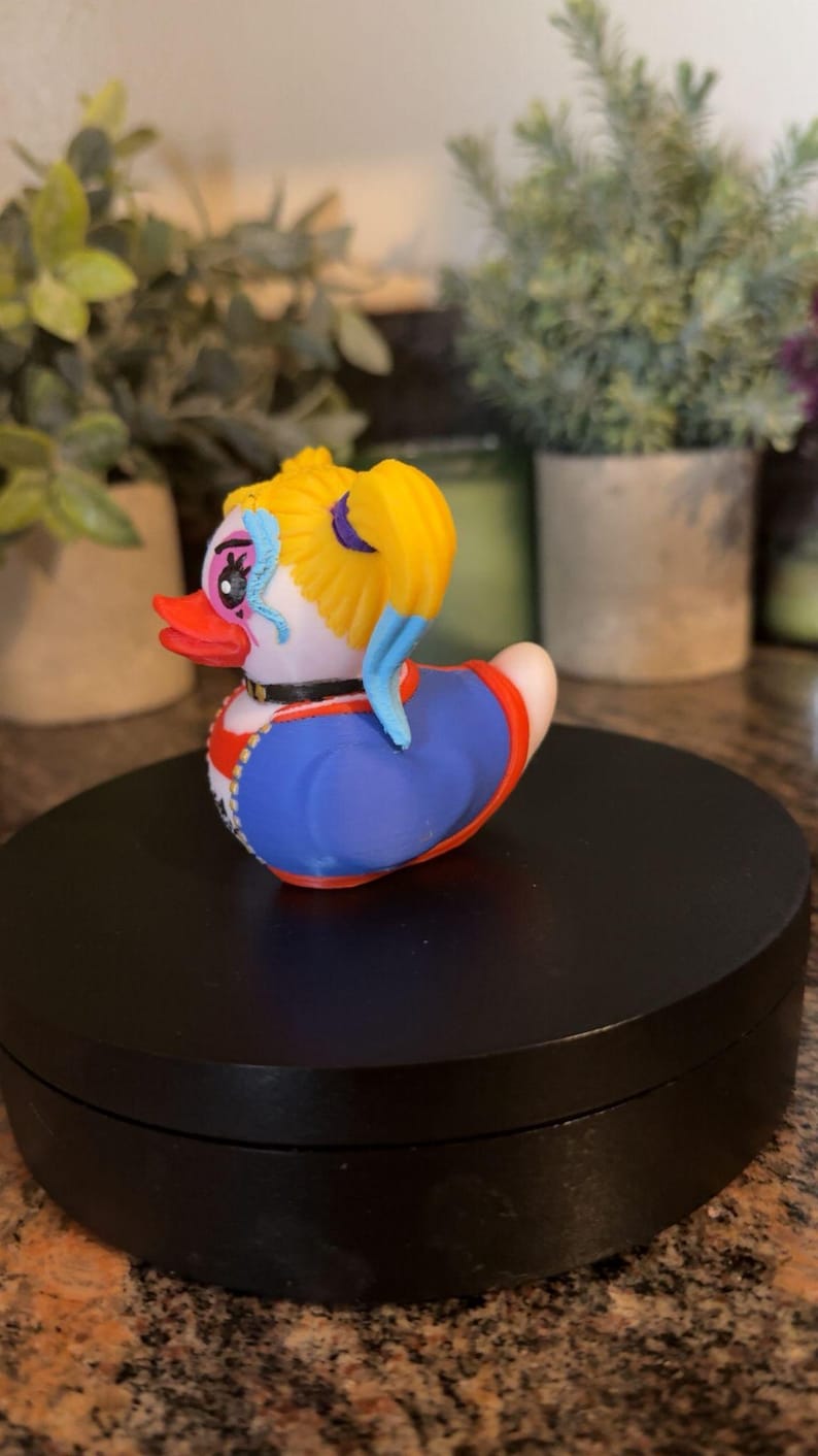 Op de afbeelding: Een decoratieve rubberen eend met een uniek ontwerp. De eend heeft een gele haarstijl, blauwe accenten en een blauwe en rode outfit. Hij heeft een zwarte kraag en een rode snavel. De eend staat op een zwart oppervlak.