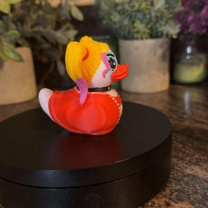Op de afbeelding: Een novelty rubberen eend met een gele paardenstaart, rode jurk en een zwarte kraag. De eend heeft een wit gezicht met roze en blauwe accenten. De eend staat op een zwart oppervlak.