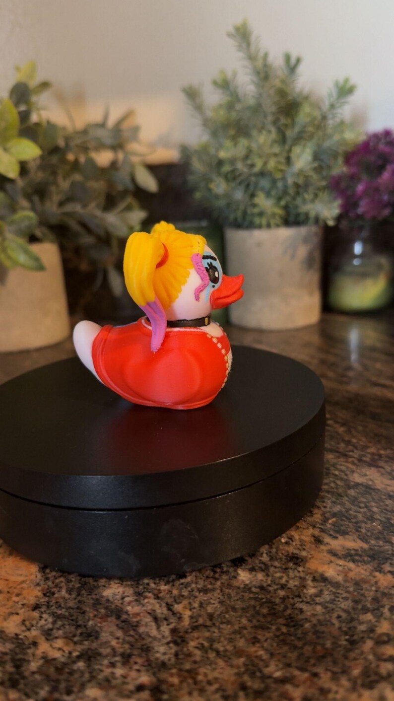 Op de afbeelding: Een novelty rubberen eend met een gele paardenstaart, rode jurk en een zwarte kraag. De eend heeft een wit gezicht met roze en blauwe accenten. De eend staat op een zwart oppervlak.