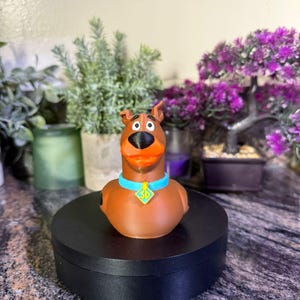 May include: A brown Scooby-Doo character figure with a black nose, orange mouth, and a light blue collar featuring a yellow diamond-shaped tag with the letters SD printed in black. The figure is shaped like a rubber duck with a smooth finish.