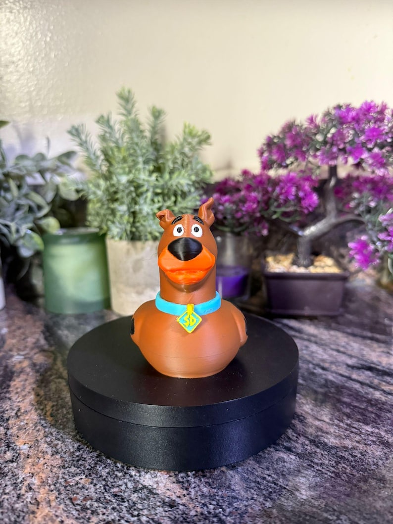 May include: A brown Scooby-Doo character figure with a black nose, orange mouth, and a light blue collar featuring a yellow diamond-shaped tag with the letters SD printed in black. The figure is shaped like a rubber duck with a smooth finish.
