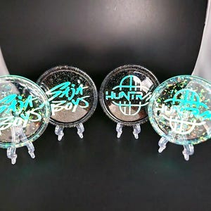 May include: Four round, clear resin coasters with glitter and teal lettering. Two coasters read "Baja Boys" and two read "HUNTR". Each coaster is displayed on a small, clear plastic stand against a black background.