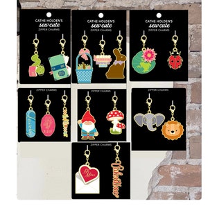 May include: A set of 12 mini zipper charms featuring various designs, including a ladybug, a bunny, a gnome, a mushroom, an elephant, a lion, a flower, a book, a basket, a frog, a heart, and an envelope. The charms are gold-colored with colorful details. The charms are packaged on a card with the text "Cathe Holden's sew cute mini zipper charms".