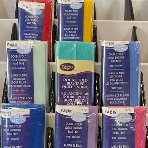 May include: A retail display of Wrights double fold quilt binding bias tape in various colors including red, yellow, light green, teal, blue, purple, and pink. Each package is labeled .875 inches or 22.2 mm and features pre-shrunk fabric.