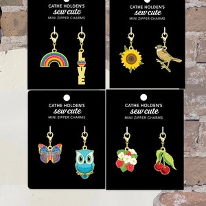 May include: A display of Cathe Holden's "sew cute" mini zipper charms. The charms include a rainbow, the word "LOVE", a sunflower with a bird, a butterfly, an owl, strawberries, and cherries. Each charm is gold-toned and attached to a black card.