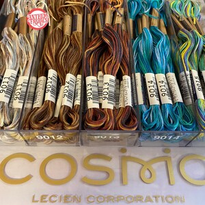 May include: A close-up of a clear plastic container filled with spools of embroidery floss. The floss is arranged in four rows, with each row containing a different color combination. The colors include shades of blue, green, brown, and beige. The spools are labeled with the numbers 9011, 9012, 9013, 9014, and 9015. The container is sitting on a white surface with the words "Cosmio Lecien Corporation" printed in gold letters.