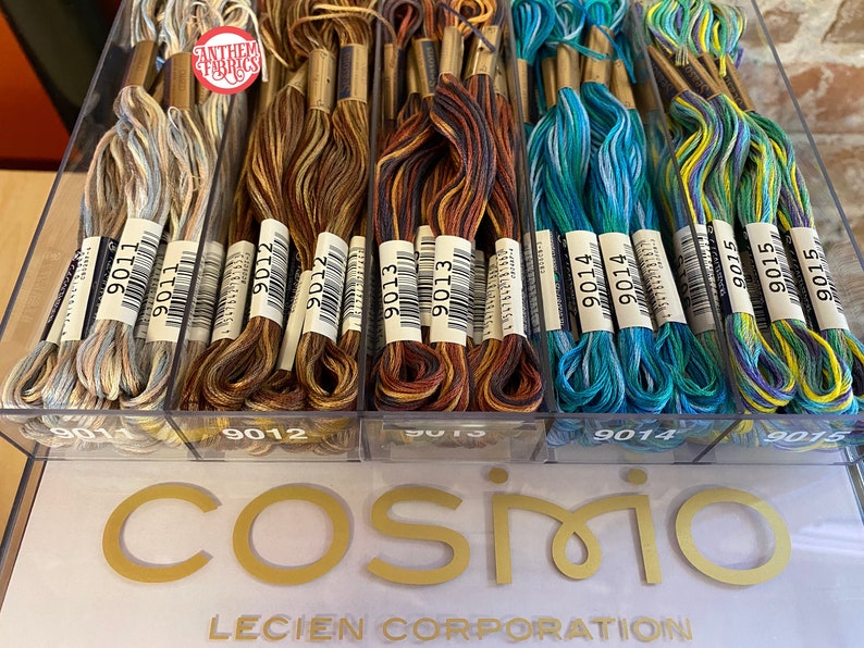May include: A close-up of a clear plastic container filled with spools of embroidery floss. The floss is arranged in four rows, with each row containing a different color combination. The colors include shades of blue, green, brown, and beige. The spools are labeled with the numbers 9011, 9012, 9013, 9014, and 9015. The container is sitting on a white surface with the words "Cosmio Lecien Corporation" printed in gold letters.