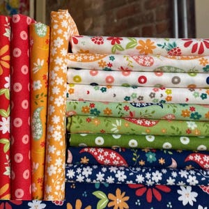 May include: A collection of folded fabric bolts and stacked fabric squares featuring various floral, paisley, and geometric patterns in a vibrant color palette of red, orange, yellow, green, and navy blue, arranged in a neat, organized display.