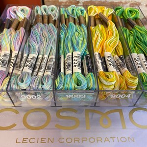 May include: A clear plastic box filled with colorful embroidery floss. The floss is organized by color and labeled with numbers. The box is labeled "COSMO LECIEN CORPORATION".