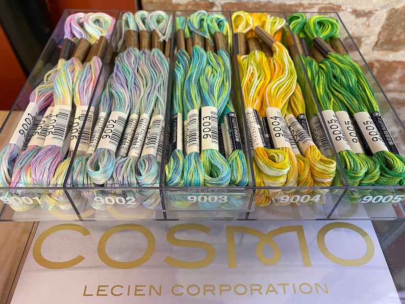 May include: A clear plastic box filled with colorful embroidery floss. The floss is organized by color and labeled with numbers. The box is labeled "COSMO LECIEN CORPORATION".