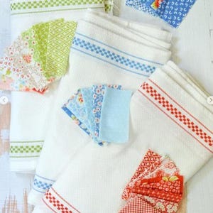 May include: White kitchen towels with decorative borders in green, blue, and red. Small fabric swatches in coordinating colors and patterns are arranged near the towels. The towels and fabric pieces are likely for home decor or crafting.