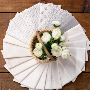 May include: A collection of white and off-white fabric squares arranged in a circular fan pattern. The fabrics feature various subtle patterns including dots, thin lines, and small floral prints. A small woven basket holds white ranunculus flowers.