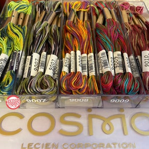 May include: A close-up of a clear plastic container filled with colorful embroidery floss. The floss is organized by color and labeled with numbers. The container is labeled with the numbers 9006, 9007, 9008, 9009, and 9010. The container is sitting on a white surface with the word "COSMIO" printed in gold letters.