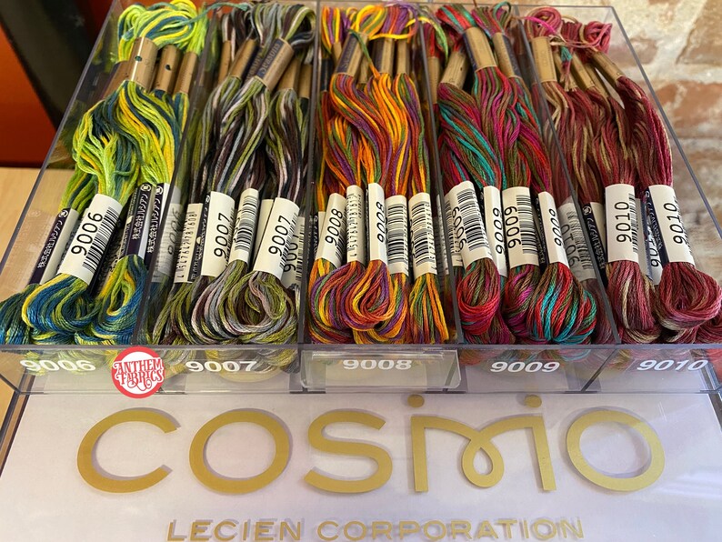 May include: A close-up of a clear plastic container filled with colorful embroidery floss. The floss is organized by color and labeled with numbers. The container is labeled with the numbers 9006, 9007, 9008, 9009, and 9010. The container is sitting on a white surface with the word "COSMIO" printed in gold letters.