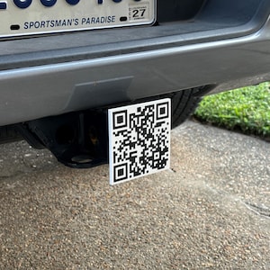 May include: A black and white QR code sticker attached to a gray vehicle's hitch receiver.