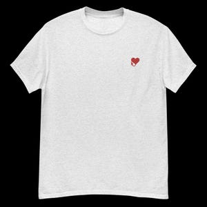 Op de afbeelding: Lichtgrijs t-shirt met een klein rood hart en duivelstaartontwerp op de linkerborst. Het shirt heeft een ronde hals en korte mouwen. De stof lijkt zacht en comfortabel.