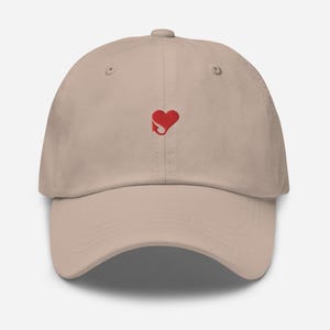 Puede incluir: Gorra de béisbol beige con visera curva. La gorra presenta un pequeño corazón rojo bordado con una cola estilizada en la parte delantera. La gorra está hecha de tela suave y tiene un diseño clásico de seis paneles.