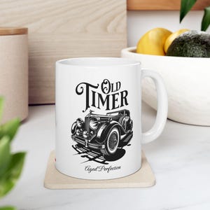 May include: White ceramic mug with a black and white vintage car design, the words "Old Timer" and "Aged Perfection" are also printed on the mug. The mug has a curved handle and is on a beige coaster.