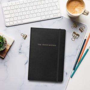Pode incluir: Um caderno preto com o texto dourado "THE TRANS AGENDA" está sobre uma superfície de mármore branco. Um teclado branco, uma chávena de café e lápis de cor também estão sobre a superfície. Também são visíveis clipes dourados e uma pequena planta.