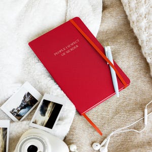 May include: A red notebook with the text "People I Suspect of Murder" on the cover. The notebook is lying on a white and brown fuzzy surface with a pen, a camera, and headphones.