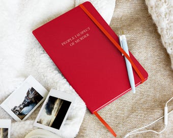 May include: A red notebook with the text "People I Suspect of Murder" on the cover. The notebook is lying on a white and brown fuzzy surface with a pen, a camera, and headphones.