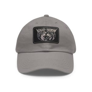 May include: A gray baseball cap with a curved brim. The cap features a rectangular patch with a black and white illustration of a snarling cat. The patch is stitched onto the front of the cap.