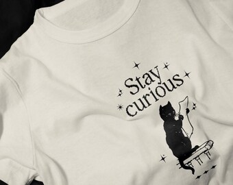 Pode incluir: Camiseta branca com um gato preto lendo um livro e o texto "Stay curious" impresso nela.