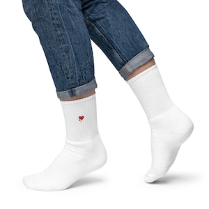 Op de afbeelding: Witte sokken met een rood hart geborduurd bij de enkel. De sokken zijn geribbeld en lijken gemaakt van een zacht, comfortabel materiaal. Gedragen met een blauwe jeans.