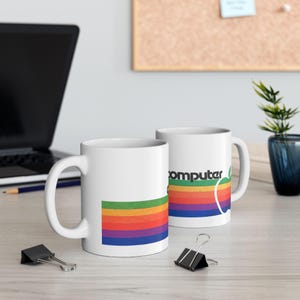 May include: Two white ceramic mugs with a rainbow stripe design and the text "computer" on one mug. The mugs are on a light wood surface.