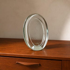May include: A clear, circular glass object with a thick, rounded frame and a slightly recessed, textured centre. The glass has a subtle green tint and a smooth, polished surface, standing upright with a flat base for stability on a flat surface.