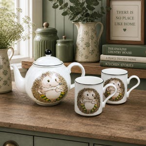 May include: A white ceramic teapot and two mugs featuring a hedgehog design. The teapot has a black rim and a black lid knob. The mugs and teapot are displayed on a wooden surface, with other decorative items in the background.
