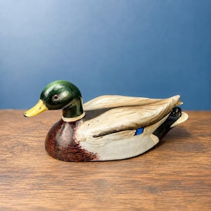 May include: A ceramic mallard duck figurine featuring a glossy green head, a bright yellow beak, a dark brown chest, and cream-coloured wings with a small blue accent patch. The duck is depicted in a resting position with its body angled to the left.
