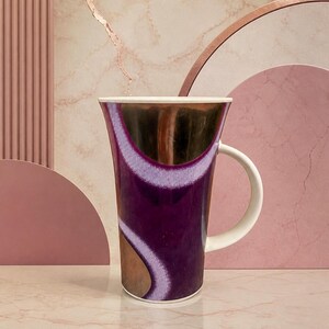 May include: A tall ceramic mug with a white handle and rim. The mug has a deep purple glaze with a gold and white abstract design. The mug is set against a pink and peach-coloured backdrop.